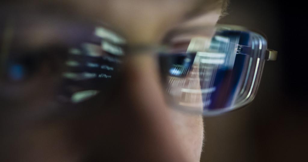 Close-up of a young programmer (18 year old male) working on a computer in a room with dimmed light. Symbols and computer program code fragments from the computer screen are reflecting in the man's glasses. High concentration. Shady atmosphere of hacking / cyber criminal.